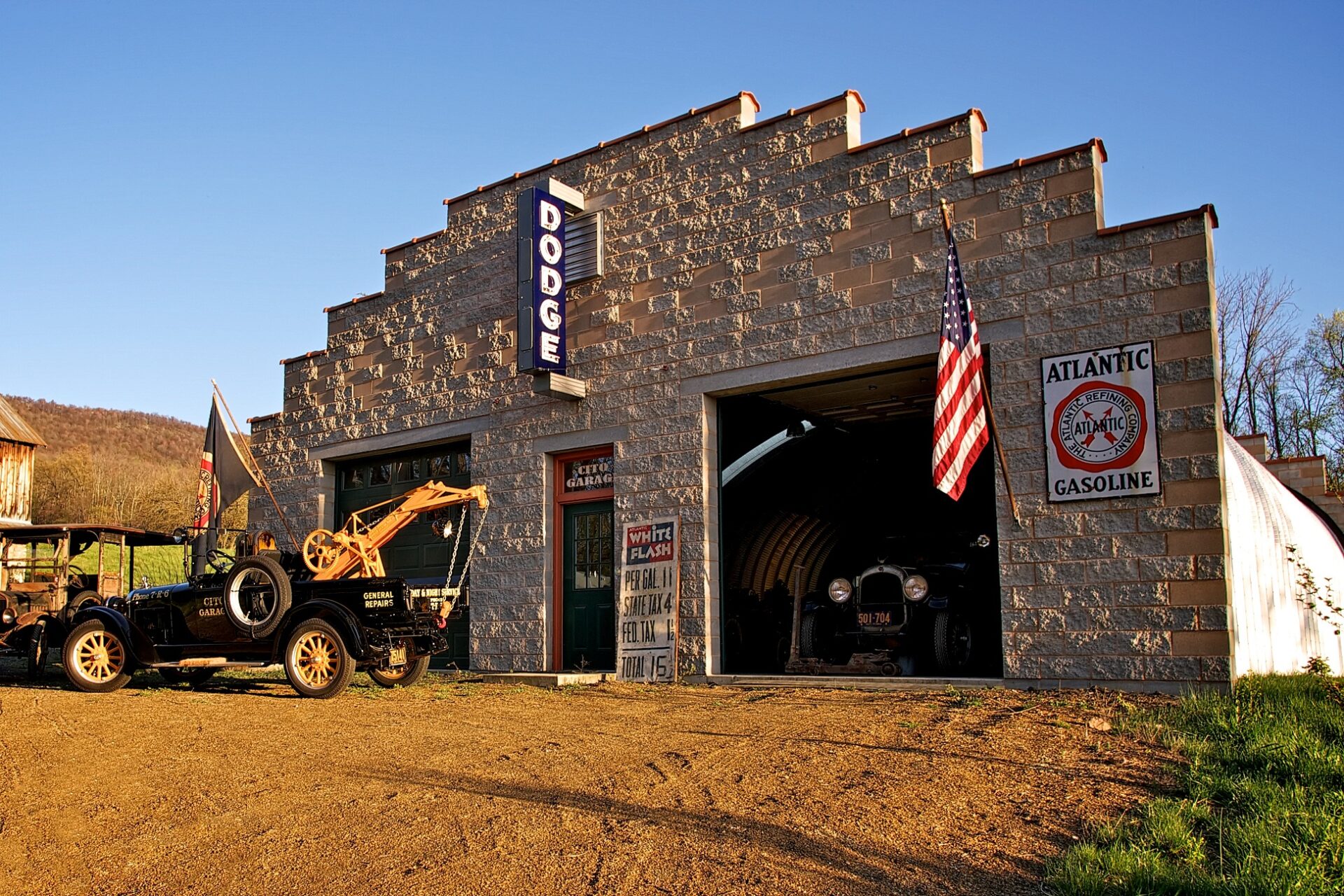 S-Model Quonset hut with custom brick end wall, an Atlantic Gasoline sign on the right, a Dodge sign in the middle, and a black vintage car on the left in front of a blue flag.