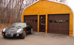 A-Model Quonset hut with a wooden end wall, brown garage doors, brown man door, and a black convertible in front of the building.